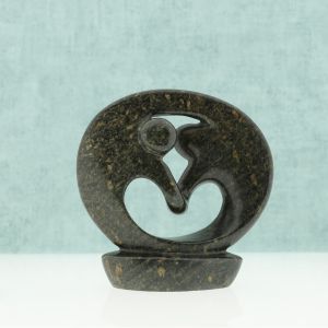 Op de foto een naturel bruinkleurig spekstenen beeld van twee figuren in een cirkelvormige omarming of dans (Ø ca. 12 cm). De vloeiende lijnen symboliseren oneindige verbinding en harmonie, u ziet zelfs een hartvorm door de stand van de armen. Vervaardigd